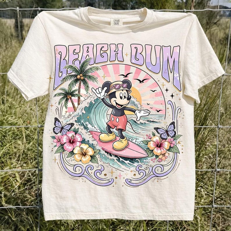 May include: Cream-colored t-shirt featuring a colorful graphic of Mickey Mouse surfing. The design includes the words "Beach Bum", palm trees, waves, butterflies, and flowers. The shirt has short sleeves and a crew neck.