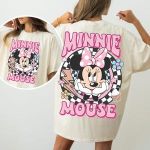 May include: Cream-colored t-shirt featuring a large graphic of Minnie Mouse with a pink bow, surrounded by flowers, stars, and the words "MINNIE MOUSE" in pink. The design is set against a black and white checkered background.