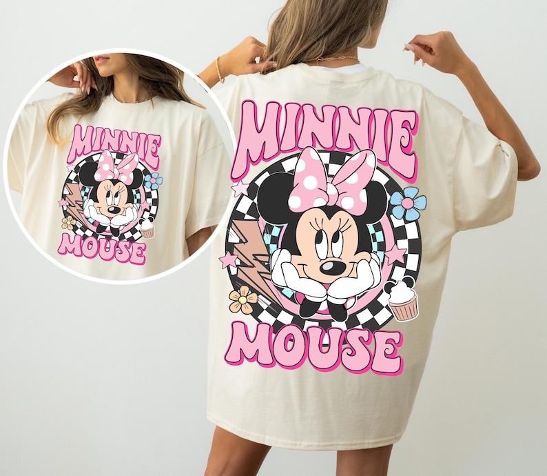 May include: Cream-colored t-shirt featuring a large graphic of Minnie Mouse with a pink bow, surrounded by flowers, stars, and the words "MINNIE MOUSE" in pink. The design is set against a black and white checkered background.