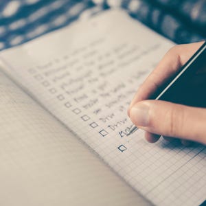May include: A close-up of a hand writing on a lined notebook with a black pen. The notebook has a grid pattern and is open to a page with a checklist. The checklist includes items such as "Plan", "Finish", "Make", "Drive", and "More".