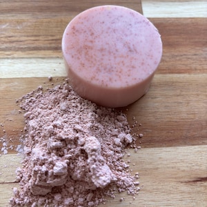 May include: A pink soap bar with a grainy texture sits on a wooden surface next to a pile of pink clay powder.