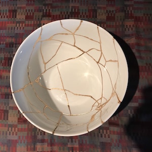 May include: A white ceramic bowl with a crackled glaze and gold accents. The bowl is cracked and repaired with gold paint.