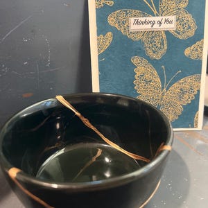 May include: A black ceramic bowl with gold-colored kintsugi repair lines. A greeting card with gold butterfly illustrations and the text "Thinking of You" is in the background.