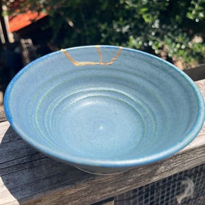 May include: A round ceramic bowl with a matte blue glaze and visible concentric ridges along the interior surface. The rim features a distinct gold-colored repair line that follows a jagged crack pattern, highlighting the kintsugi technique.