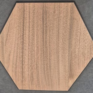 May include: A hexagonal wooden tile with a light brown wood grain pattern. The tile has a dark border and is set against a grey background. The wood grain runs vertically across the surface.