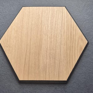 Könnte beinhalten: Ein sechseckiges Holzbrett mit einem hellbraunen Holzmaserungsmuster. Das Brett hat einen schwarzen Rand und ist vor einem dunkelgrauen Hintergrund platziert. Die Holzmaserung verläuft vertikal.