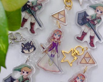 May include: Acrylic keychains featuring characters from a video game. The keychains include images of a character in a green tunic and a princess in a purple dress, along with star and triangle charms. The keychains have silver and gold hardware.