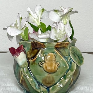May include: A ceramic vase with a frog design, holding white flowers with green stems. The vase is green, brown, and tan, with a frog on a lily pad. A red flower is on the side of the vase.