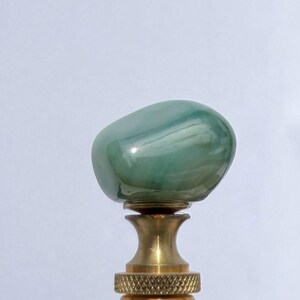 May include: A lamp finial featuring a smooth, polished, rounded green stone top with swirling white inclusions, mounted on a tiered, knurled brass base with a threaded metal screw at the bottom for secure attachment to a lamp harp.