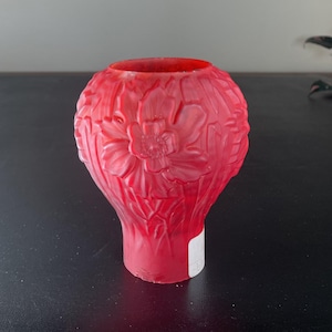 May include: A red glass vase with a floral design. The vase has a bulbous top and a narrower base. The floral pattern is embossed on the surface of the vase. The vase is made of red glass.