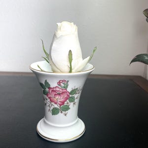 May include: A white ceramic vase with a pink and green floral design, holding a white rose. The vase has a gold trim and a flared top. The rose is in full bloom, with green stems and leaves.
