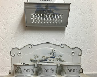 May include: White enamel kitchen shelf with three small white bowls labeled "Seife", "Soda", and "Sand". The shelf has a decorative windmill design and is mounted on a white tiled wall. Above the shelf is a white enamel box with a cut-out design and the word "Zwiebeln" written on it.
