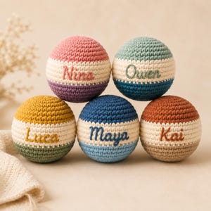 Peut inclure: Cinq balles en crochet sphériques disposées en pyramide, chacune présentant une bande centrale de couleur crème brodée avec les prénoms Nina, Owen, Luca, Maya et Kai. Les balles sont réalisées dans diverses couleurs dont rose, violet, vert, bleu et orange.