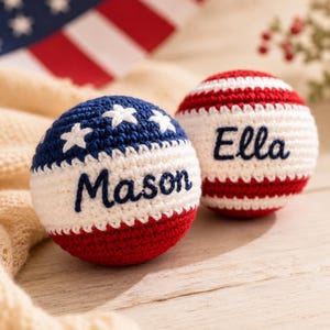 May include: Two round, crocheted yarn balls featuring red, white, and blue patterns. One ball has a blue top with white stars and the name Mason in blue script. The other ball has red and white stripes with the name Ella in blue script.