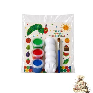 May include: A clear plastic bag contains a children's craft kit. The kit includes a colorful illustration of a caterpillar, paint pots in green, red, and blue, white caterpillar-shaped pieces, and a paintbrush. The text "THE VERY HUNGRY CATERPILLAR" is visible.