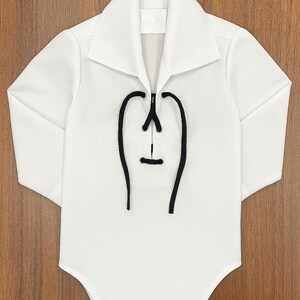 May include: White baby bodysuit with long sleeves and a collared neckline. The front features a black decorative design. The bodysuit has three snap closures at the bottom. The garment is made of a soft, comfortable material.