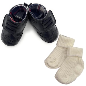 May include: A pair of black baby shoes with a classic brogue design, paired with two cream-colored ribbed socks. The shoes have a plaid lining and are designed to look like miniature dress shoes.