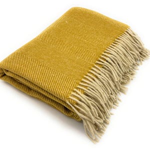 May include: Mustard yellow throw blanket with a herringbone pattern and fringed edges. The folded blanket is made of a soft, textured material. The fringe is a light beige color.