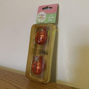 May include: A package containing two ladybug knobs. The knobs are red with orange spots and are packaged in a clear plastic container. The package has a pink and green label that reads "Restore, RESTYLE KIDS, Set of 2, Lady Bug Knobs."