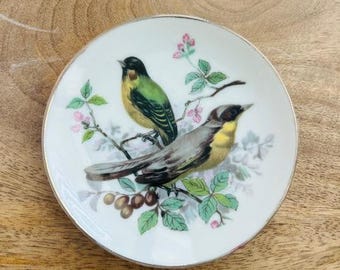 May include: A white porcelain plate with a gold rim features a hand-painted design of two birds perched on a branch with green leaves and pink flowers.