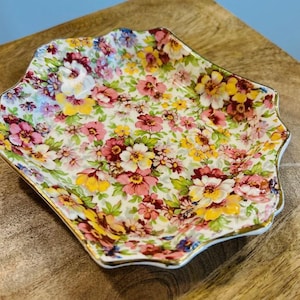 May include: A small, square, white ceramic dish with a scalloped edge. The dish is decorated with a floral pattern in shades of pink, yellow, and green.