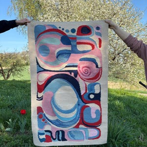 May include: A rectangular tapestry with an abstract design in shades of pink, blue, and red on a beige background. The artwork features organic shapes and curves. The tapestry is held up in a grassy outdoor setting.