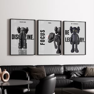 May include: Three framed art prints featuring black and gray figures with text. The first print says "DISCIPLINE." The second print says "FOCUS." The third print says "BE LEGENDARY." The prints are displayed above a black leather sofa.