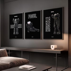 May include: Three framed black art prints with white text and black figures. The prints read "DISCIPLINE", "FOCUS", and "HUSTLE". Each print features a different pose of a black figure with white eyes. The prints are displayed on a dark grey wall.