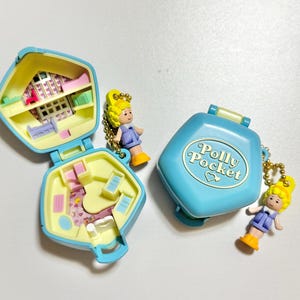 Może przedstawiać: Trzy zabytkowe kompakty Polly Pocket w kolorze niebieskim i ż&oacute;łtym, z miniaturowymi figurkami. Jeden kompakt jest otwarty, odsłaniając wnętrze domku dla lalek. Zamknięty kompakt ma napis "Polly Pocket". Figurki mają ż&oacute;łte włosy i noszą fioletowe sukienki.