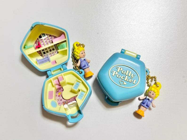 Może przedstawiać: Trzy zabytkowe kompakty Polly Pocket w kolorze niebieskim i ż&oacute;łtym, z miniaturowymi figurkami. Jeden kompakt jest otwarty, odsłaniając wnętrze domku dla lalek. Zamknięty kompakt ma napis "Polly Pocket". Figurki mają ż&oacute;łte włosy i noszą fioletowe sukienki.