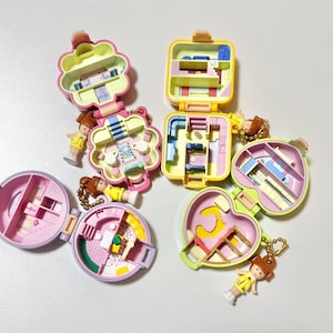 May include: A collection of vintage miniature playsets in various shapes, including a flower, heart, and square. Each playset is open, revealing colorful interior designs and small figurines. The toys are in shades of pink, yellow, and green.