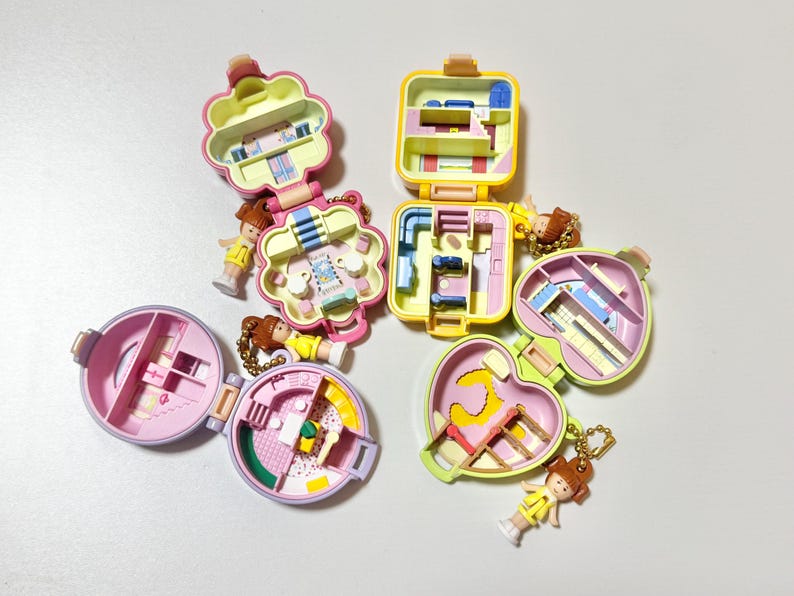 May include: A collection of vintage miniature playsets in various shapes, including a flower, heart, and square. Each playset is open, revealing colorful interior designs and small figurines. The toys are in shades of pink, yellow, and green.
