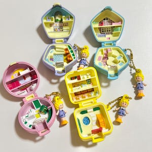 Może przedstawiać: Kolekcja miniaturowych zestaw&oacute;w do zabawy w stylu vintage w r&oacute;żnych pastelowych kolorach, w tym r&oacute;żowym, ż&oacute;łtym i niebieskim. Każdy zestaw otwiera się, ukazując szczeg&oacute;łowe wnętrze z maleńkimi meblami i akcesoriami. Do zestaw&oacute;w dołączone są małe blond włosy.