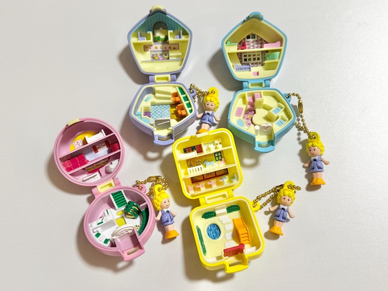 Może przedstawiać: Kolekcja miniaturowych zestaw&oacute;w do zabawy w stylu vintage w r&oacute;żnych pastelowych kolorach, w tym r&oacute;żowym, ż&oacute;łtym i niebieskim. Każdy zestaw otwiera się, ukazując szczeg&oacute;łowe wnętrze z maleńkimi meblami i akcesoriami. Do zestaw&oacute;w dołączone są małe blond włosy.