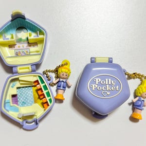 Może przedstawiać: Dwa kompaktowe Polly Pocket w kolorze lawendowym i ż&oacute;łtym. Jeden jest otwarty, ukazując miniaturowe wnętrze z meblami i akcesoriami. Drugi jest zamknięty, z napisem "Polly Pocket". Każdy ma małą lalkę.