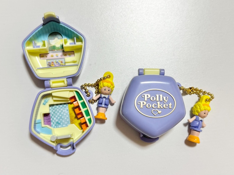 Może przedstawiać: Dwa kompaktowe Polly Pocket w kolorze lawendowym i ż&oacute;łtym. Jeden jest otwarty, ukazując miniaturowe wnętrze z meblami i akcesoriami. Drugi jest zamknięty, z napisem "Polly Pocket". Każdy ma małą lalkę.
