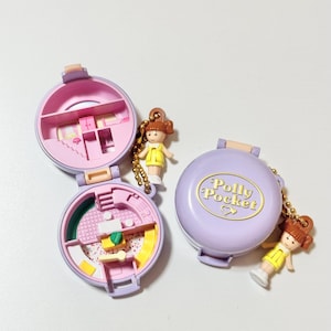 May include: Two vintage Polly Pocket compacts, one open and one closed, in shades of pink and purple. The open compacts reveal miniature rooms. Each compact has a small doll charm attached. The closed compact has the words "Polly Pocket" in gold.