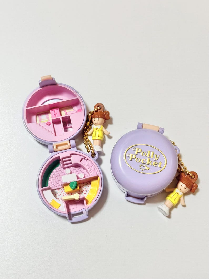 May include: Two vintage Polly Pocket compacts, one open and one closed, in shades of pink and purple. The open compacts reveal miniature rooms. Each compact has a small doll charm attached. The closed compact has the words "Polly Pocket" in gold.