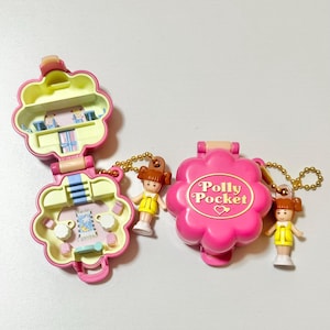 May include: Two vintage Polly Pocket compacts in pink, shaped like flowers, with miniature figures. One is open, revealing a small interior scene. The other is closed, with the words "Polly Pocket" on the front. Each has a gold chain.