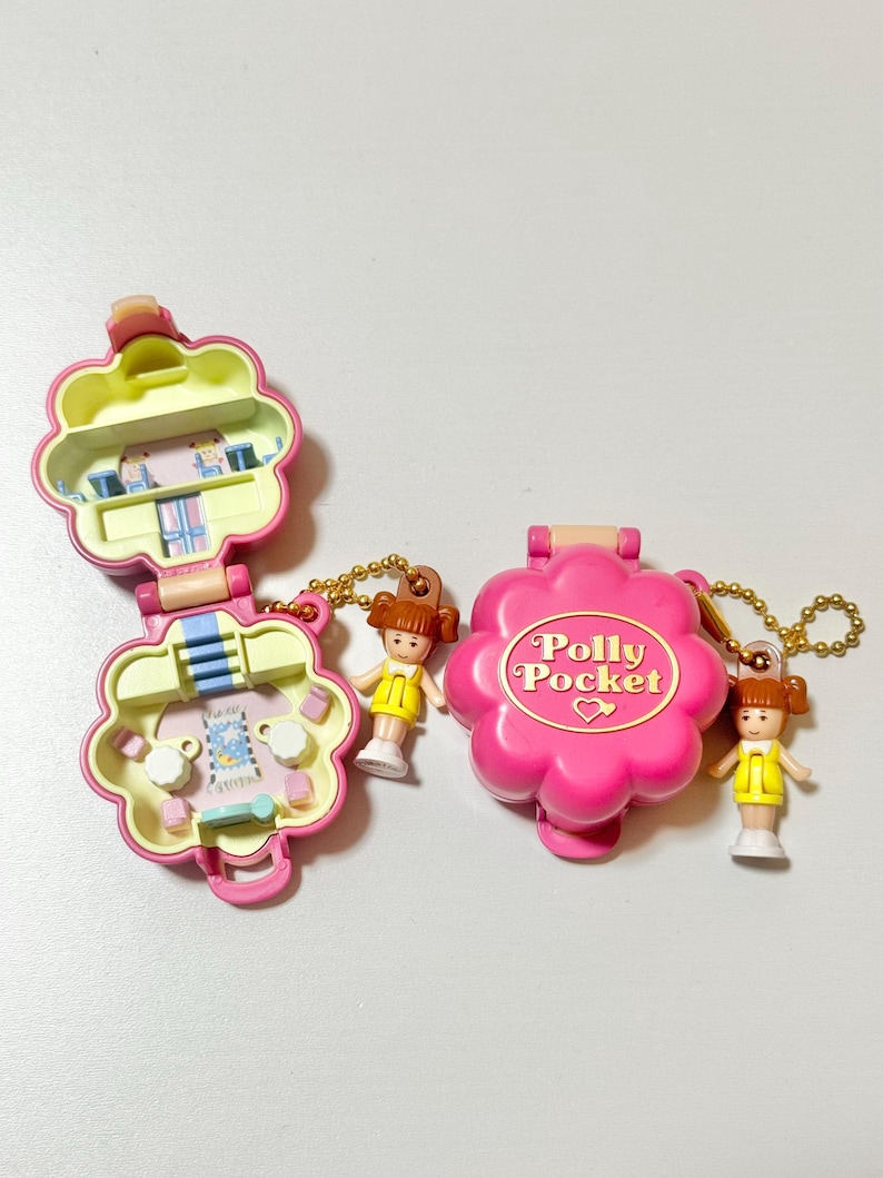 May include: Two vintage Polly Pocket compacts in pink, shaped like flowers, with miniature figures. One is open, revealing a small interior scene. The other is closed, with the words "Polly Pocket" on the front. Each has a gold chain.