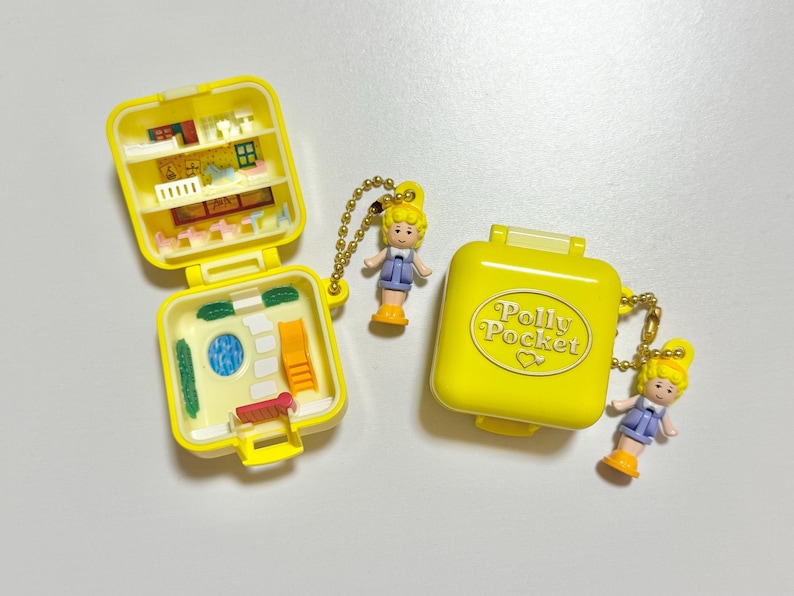 Może przedstawiać: Dwa ż&oacute;łte kompakty Polly Pocket, jeden otwarty, aby odsłonić miniaturowe wnętrze z meblami i lalką. Drugi kompakt jest zamknięty ze słowami "Polly Pocket". Każdy ma dołączoną lalkę.