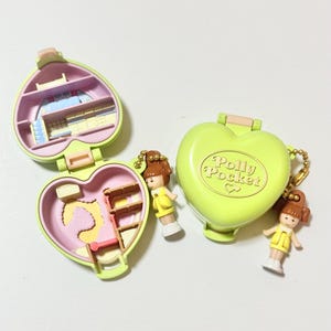 May include: Two vintage Polly Pocket compacts in the shape of hearts, one open and one closed. The open compacts reveal miniature rooms with furniture. Two small dolls with brown hair and yellow outfits are attached to the compacts.