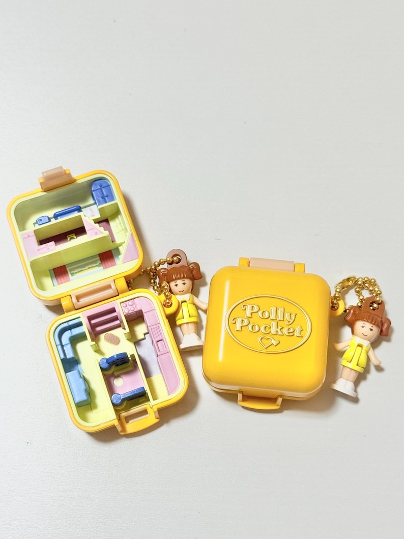 May include: Yellow Polly Pocket compact with the text "Polly Pocket" and two miniature figures. Two open compacts reveal interior rooms. The figures are wearing yellow dresses. The compacts are on a white surface.