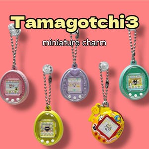 以下が含まれることがあります： ピンク、紫、緑、黄色、黄色の5つのTamagotchi3ミニチュアチャーム。各チャームには、ピクセル化された画像を表示する小さな画面があり、シルバーチェーンから吊り下げられています。「Tamagotchi3 miniature charm」の文字が上部にあります。