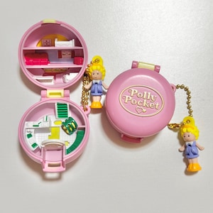 Może przedstawiać: Zestaw do zabawy Polly Pocket w stylu vintage, z r&oacute;żowym kompaktem z zapięciem w kształcie serca. Otwarty kompakt odsłania miniaturowe wnętrze z meblami i małą lalką. Dwie lalki są przymocowane złotymi łańcuszkami.