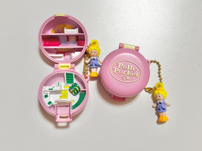 Może przedstawiać: Zestaw do zabawy Polly Pocket w stylu vintage, z r&oacute;żowym kompaktem z zapięciem w kształcie serca. Otwarty kompakt odsłania miniaturowe wnętrze z meblami i małą lalką. Dwie lalki są przymocowane złotymi łańcuszkami.