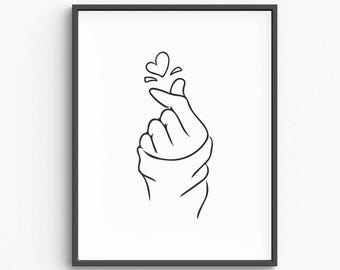 Op de afbeelding: Zwart ingelijste kunstdruk. Een minimalistische lijntekening toont een hand die een hart vormt met de vingers, met een klein hartje erboven. Het kunstwerk staat op een witte achtergrond.