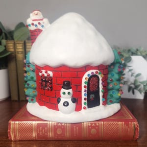 May include: A ceramic Christmas house with a red brick facade, a white snow-covered roof, and a chimney with Santa Claus. The house features a snowman, a decorated door, and Christmas tree accents. It sits atop a red book.