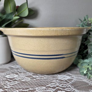 May include: A tan stoneware mixing bowl with a speckled texture and two horizontal dark blue stripes encircling the exterior. The bowl features a wide, flared rim and small side handles, providing a functional design for kitchen use.