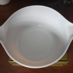 May include: A white, vintage Pyrex mixing bowl with two handles. The bowl has a smooth, glossy finish and a rounded shape. The bowl is sitting on a gold-colored surface.